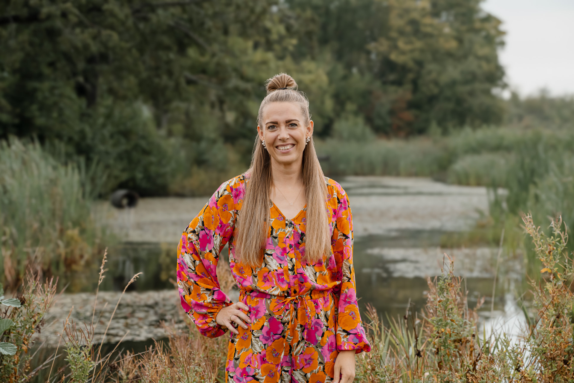 Nicole Mørck i blomstret kjole smilende ved en sø omgivet af højt græs og træer