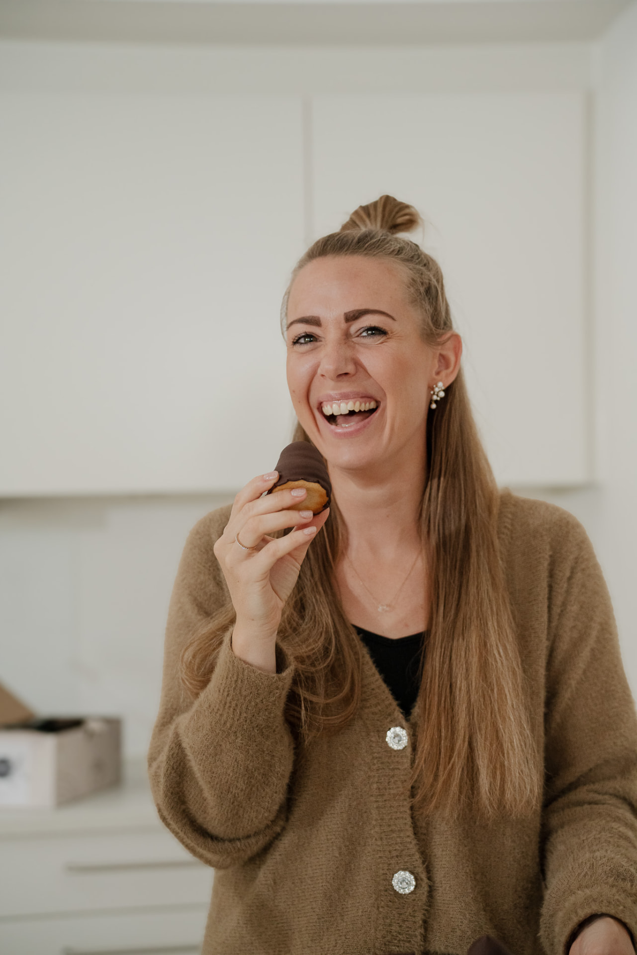 Nicole griner højlydt med en chokoladeovertrukket kage i hånden i sit lyse køkken