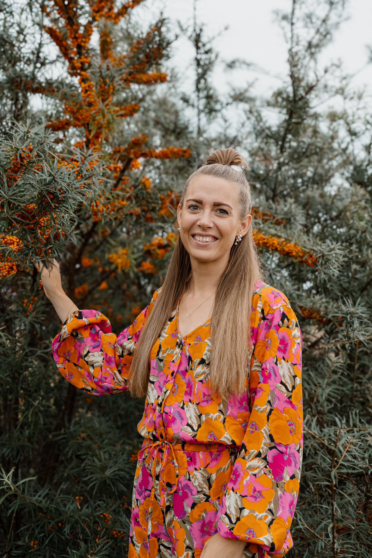 Nicole i blomstret kjole står ved en havtornbusk med orange bær og smiler